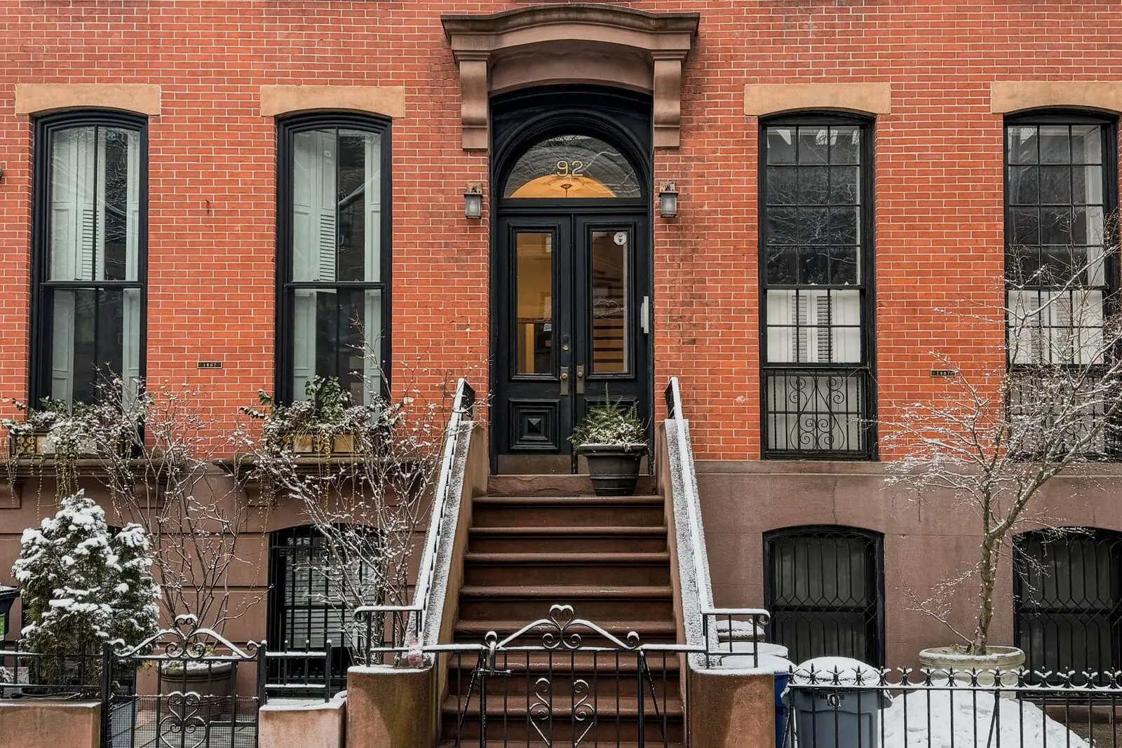 A red-brick New York house with a black front door at the top of a stoop, light snow on the railings, planters and sidewalk.