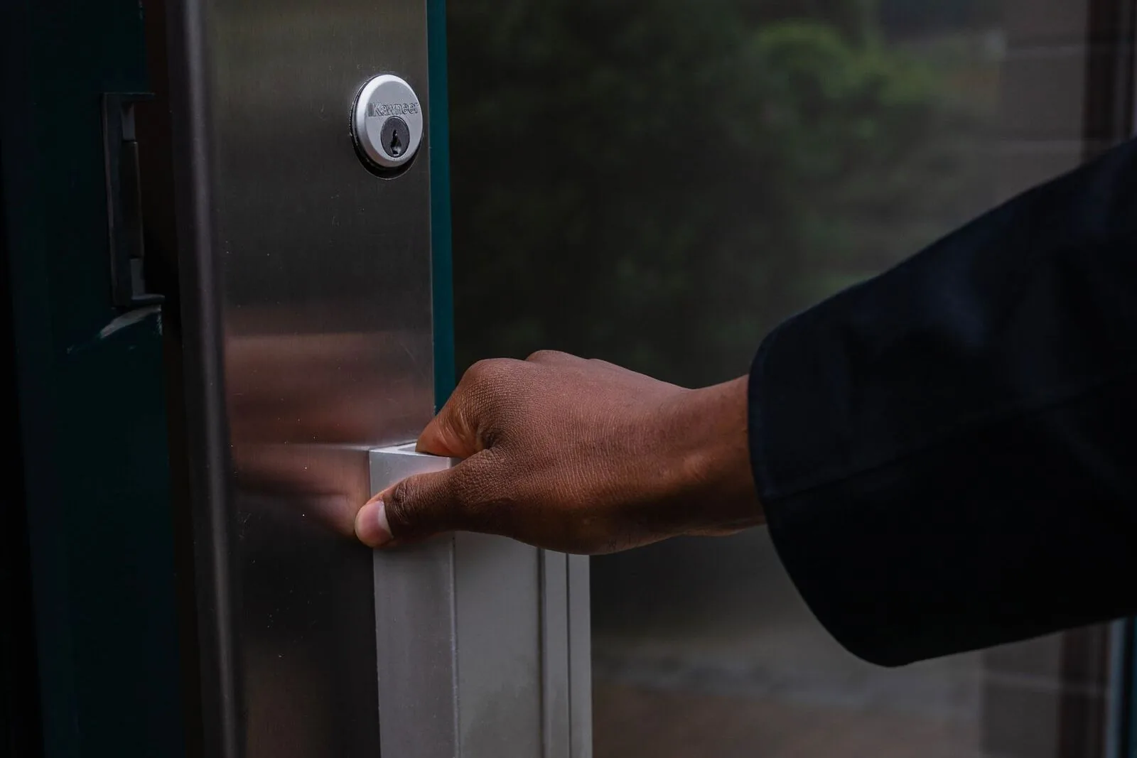 A hand on the push handle of a commercial glass door, with a keyed cylinder lock set into the metal frame above it.
