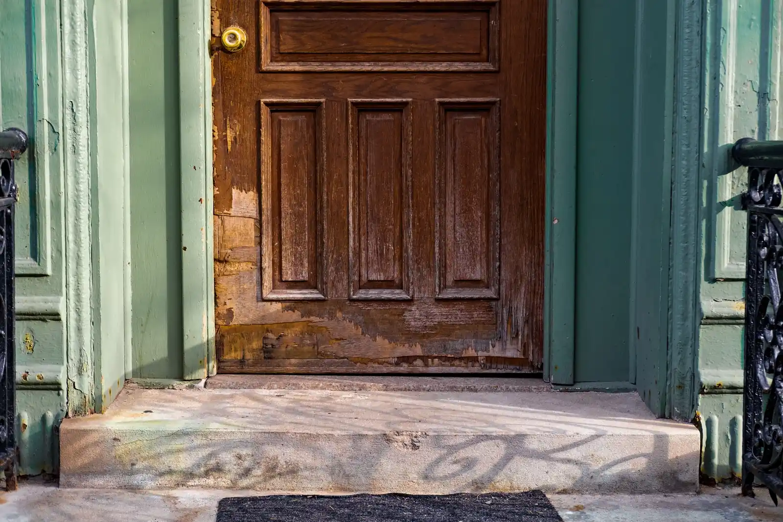 A brown paneled front door with a brass knob and a scraped, battered lower edge, between green door frames above a stone step.