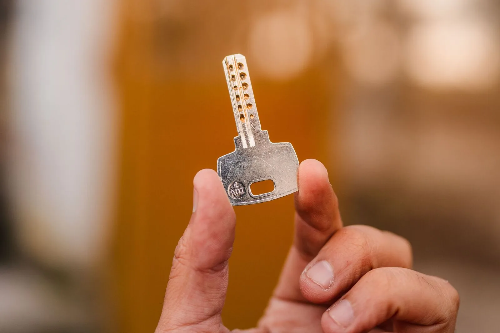 A hand holding up a single dimple-style security key against a blurred amber and off-white background.