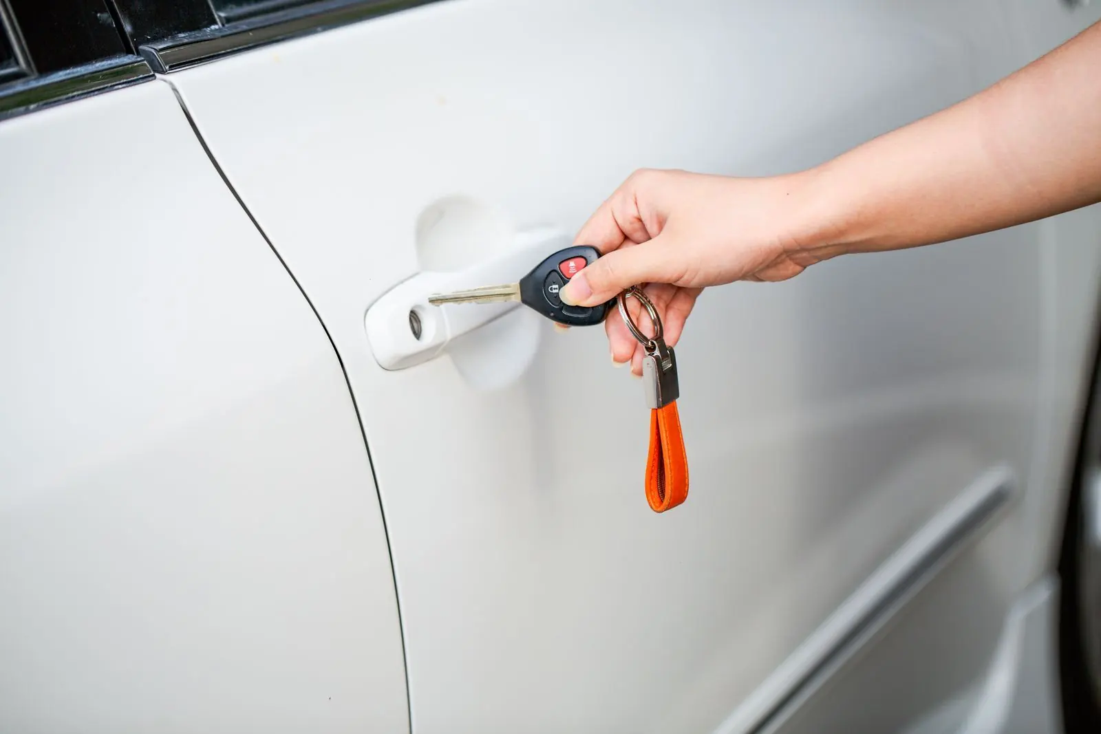 A hand turning a metal key with a black remote head in the door lock of a white car, an orange strap hanging from the ring.
