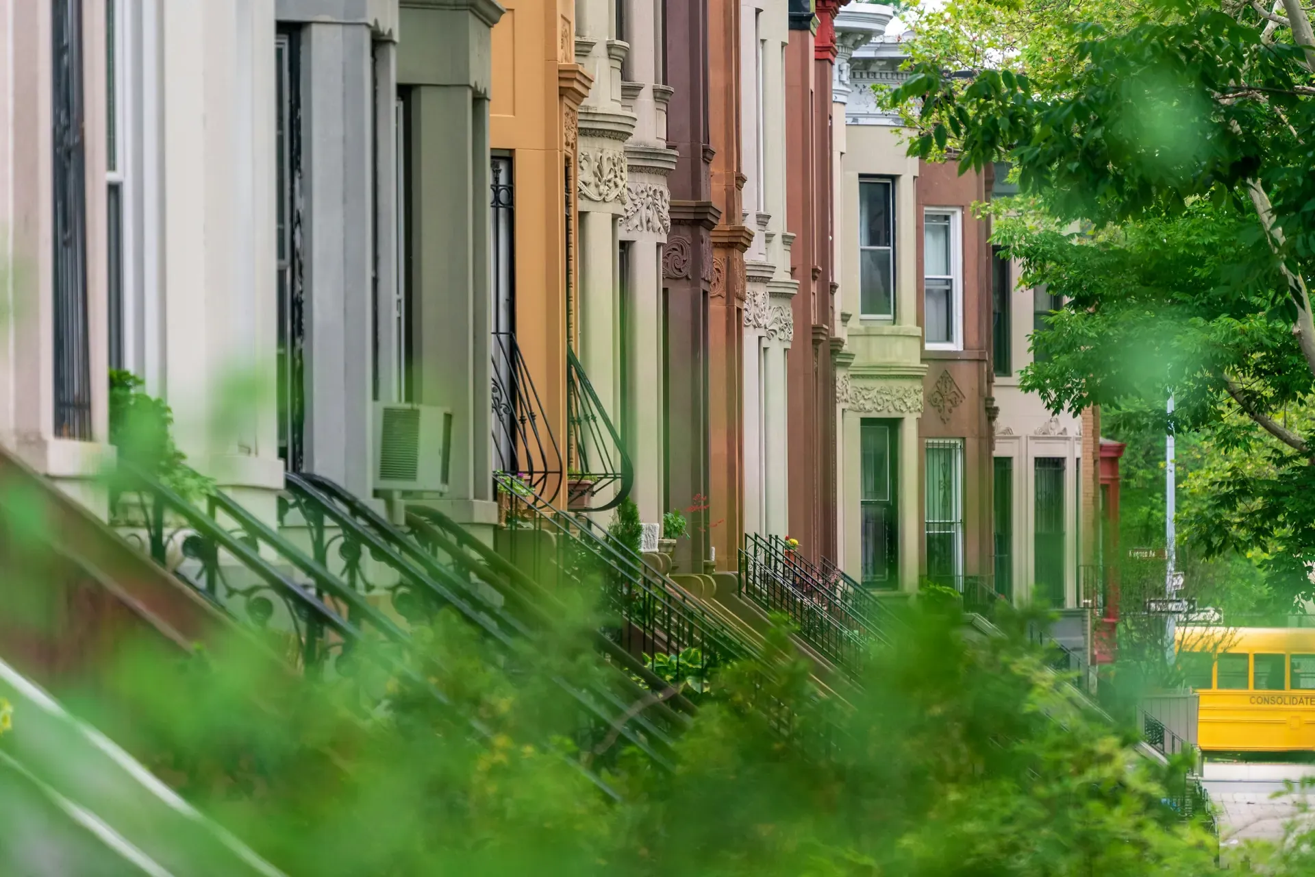 Tree-lined Brooklyn row houses with iron stoop railings. A yellow school bus is visible in the distance.