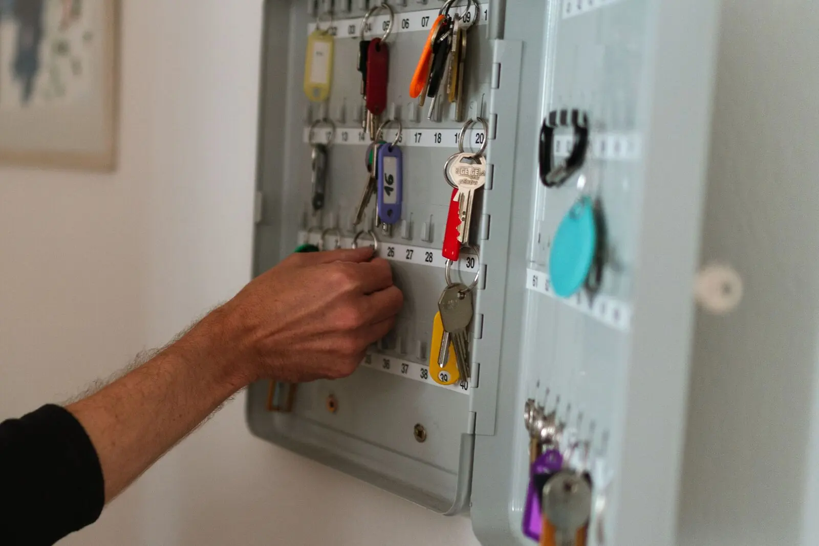 A hand reaching into an open wall-mounted key cabinet with numbered hooks and color-tagged keys.