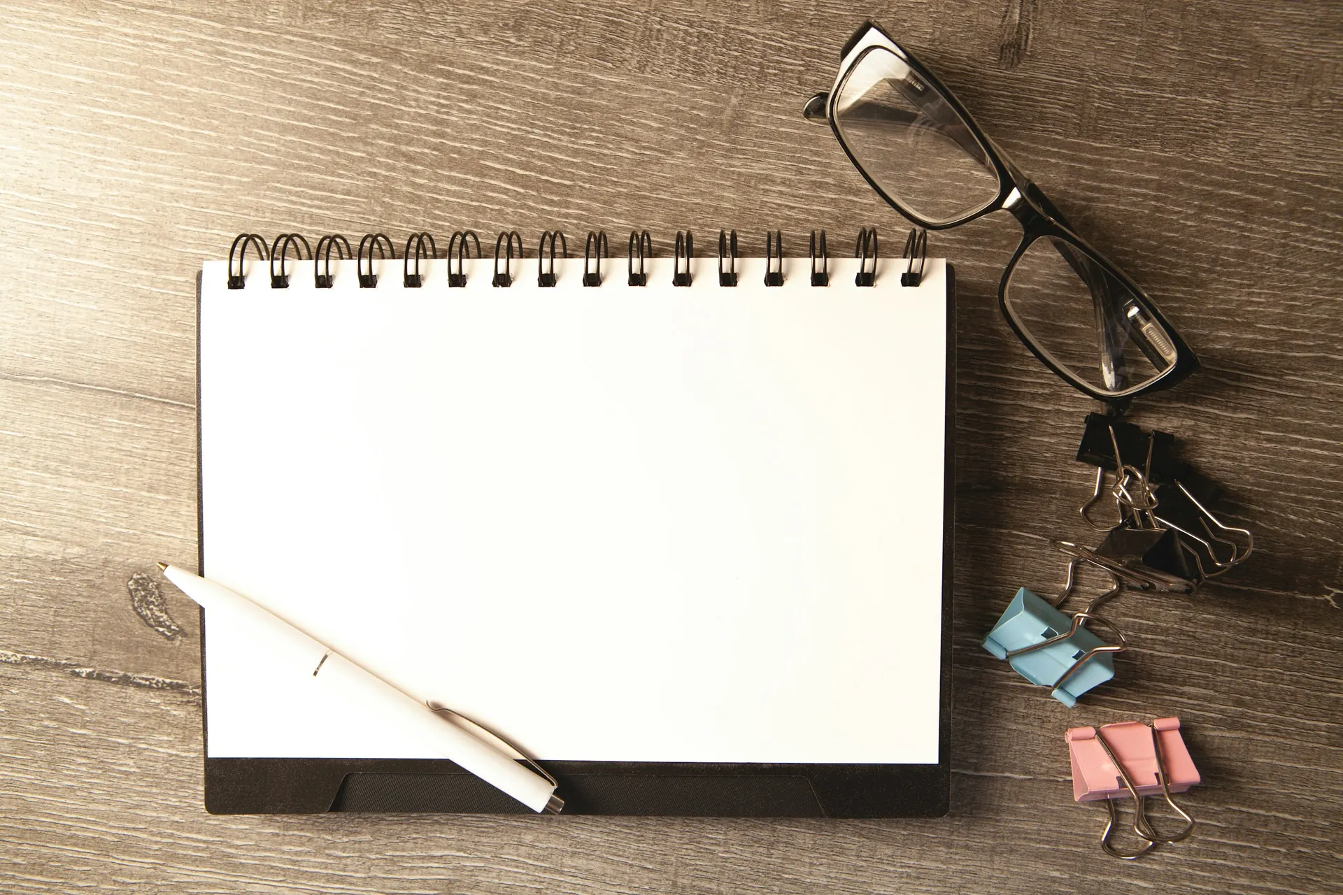 A blank spiral notebook rests on a wooden desk. Glasses, a white pen and colored binder clips sit beside it.