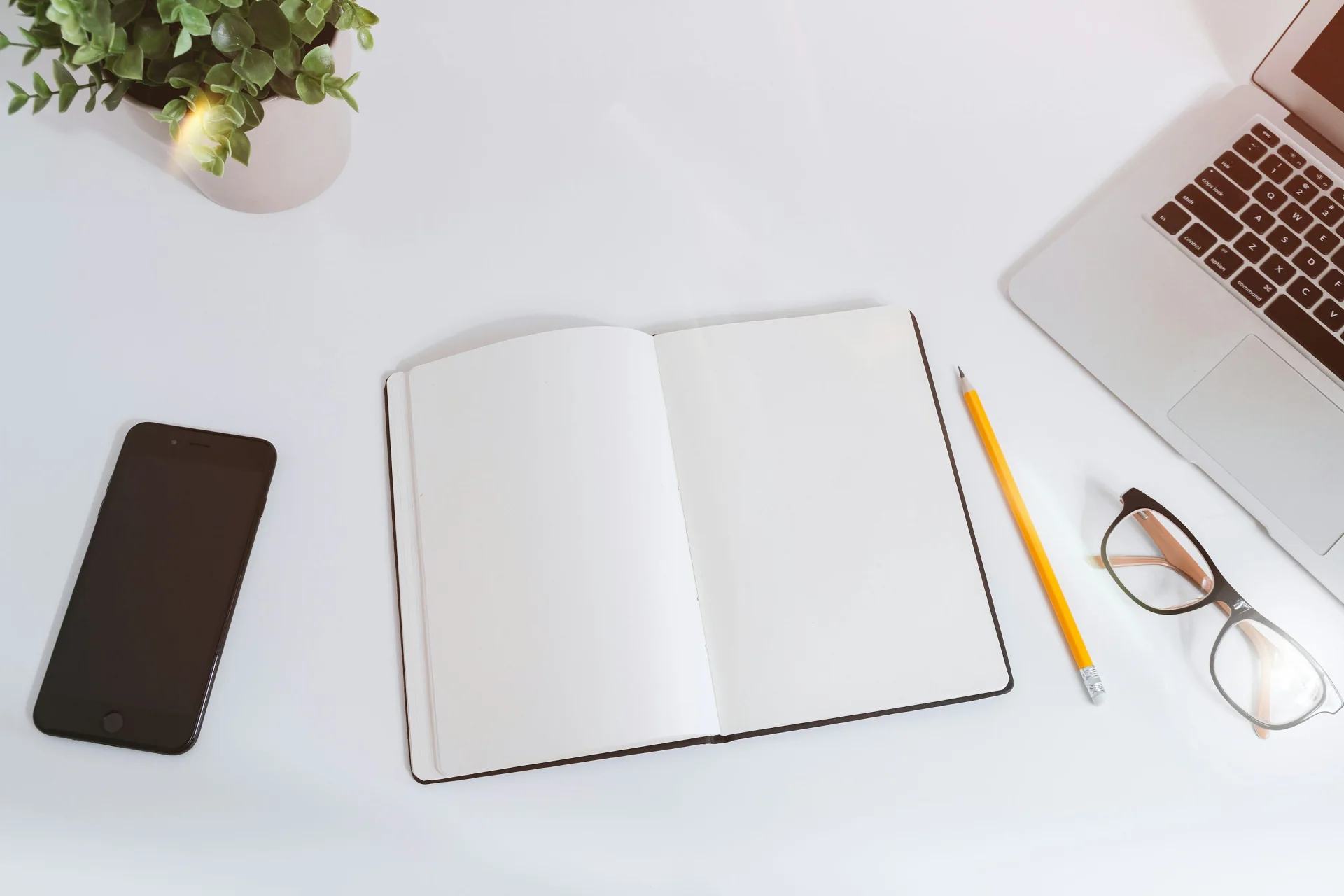 Open blank notebook beside a phone and laptop on a white desk