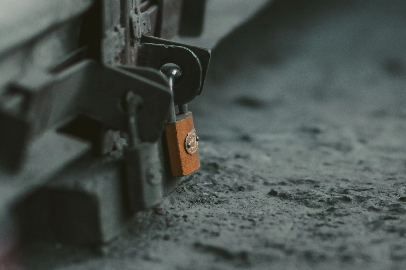Close-up of a small brass padlock hanging from the hasp at the bottom corner of a grey roll-down metal shutter, with dusty concrete behind it.