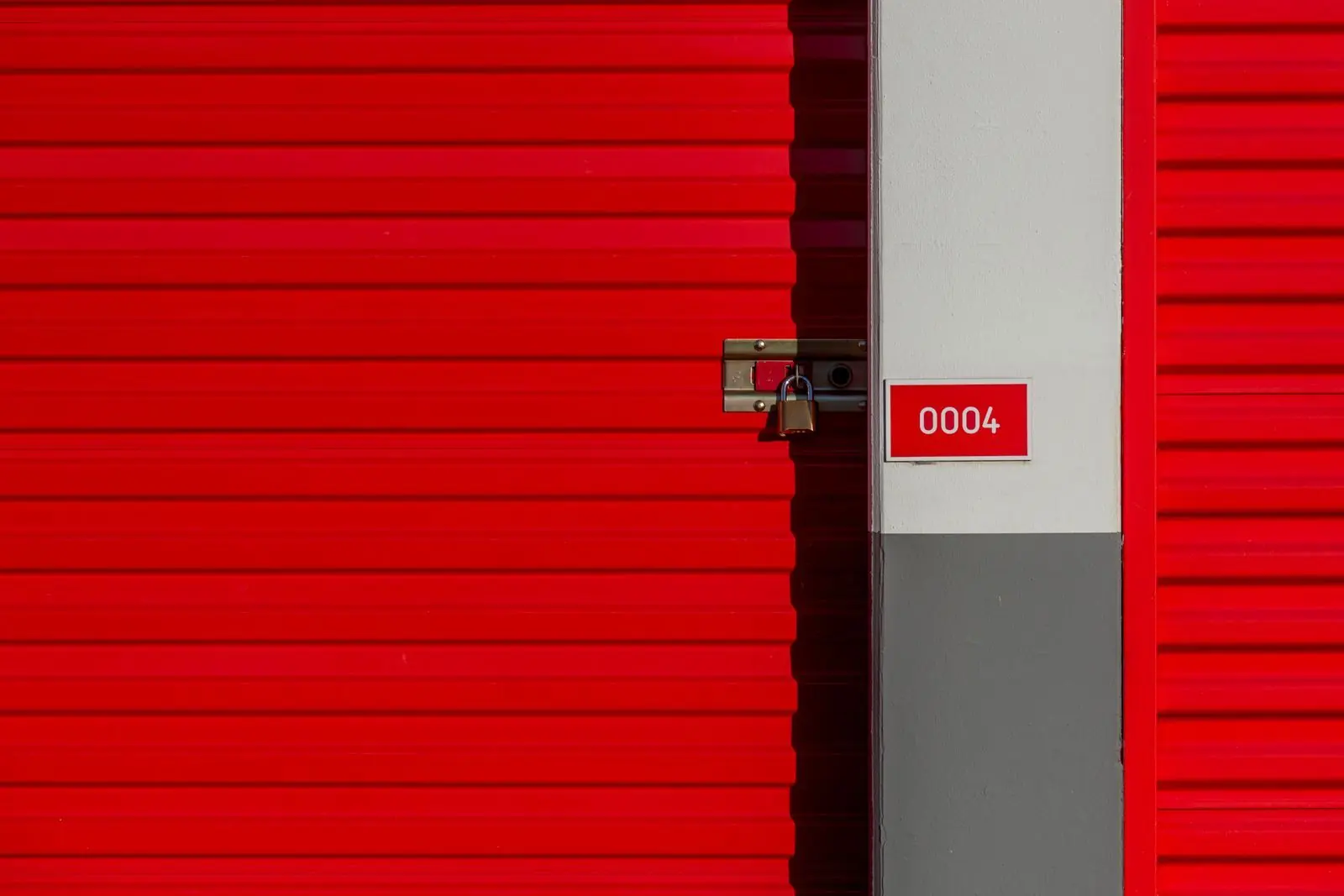 A red roll-up storage unit door closed with a brass padlock, beside a unit number sign.