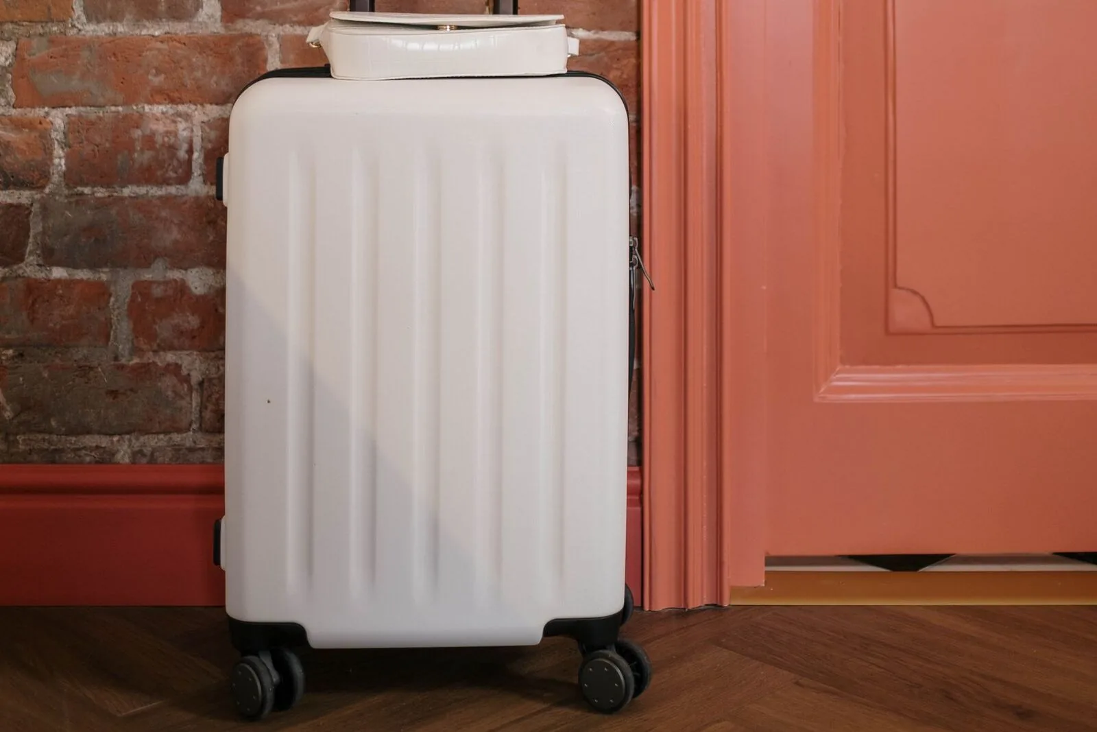 A white hard-shell suitcase standing on a wooden floor beside a closed coral-colored door and a brick wall.
