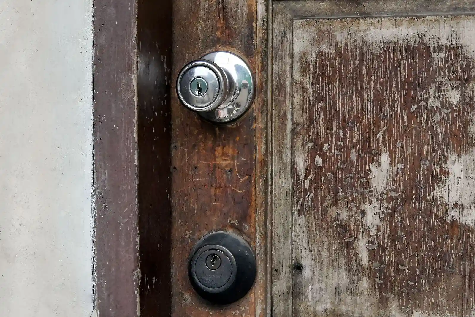 A weathered brown wooden door with a chrome keyed knob above a separate black keyed deadbolt.